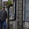 Commuter-Gloves-and-Furnace-Flat-Cap-1-3.jpg A man wearing a flat cap, checked shirt and dark puffer vest walks with his hands in his pockets down a narrow lane of old stone buildings adorned with hanging baskets and large mullioned windows.