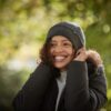 Contour-Eco-Polylana-Beanie-scaled-2.jpg Smiling woman adjusting a grey knitted beanie, wearing a black puffer jacket with curly hair peeking out against a sunlit green bokeh background.