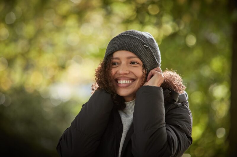 Contour-Eco-Polylana-Beanie-scaled-2.jpg Smiling woman adjusting a grey knitted beanie, wearing a black puffer jacket with curly hair peeking out against a sunlit green bokeh background.