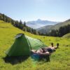 Coshee-2-Lifestyle-1500x1500-1-3.jpg Person lying on their back with hands behind their head beside a bright green tent on a sunlit grassy slope, with tree-lined hills and distant snow-capped mountains under a clear blue sky