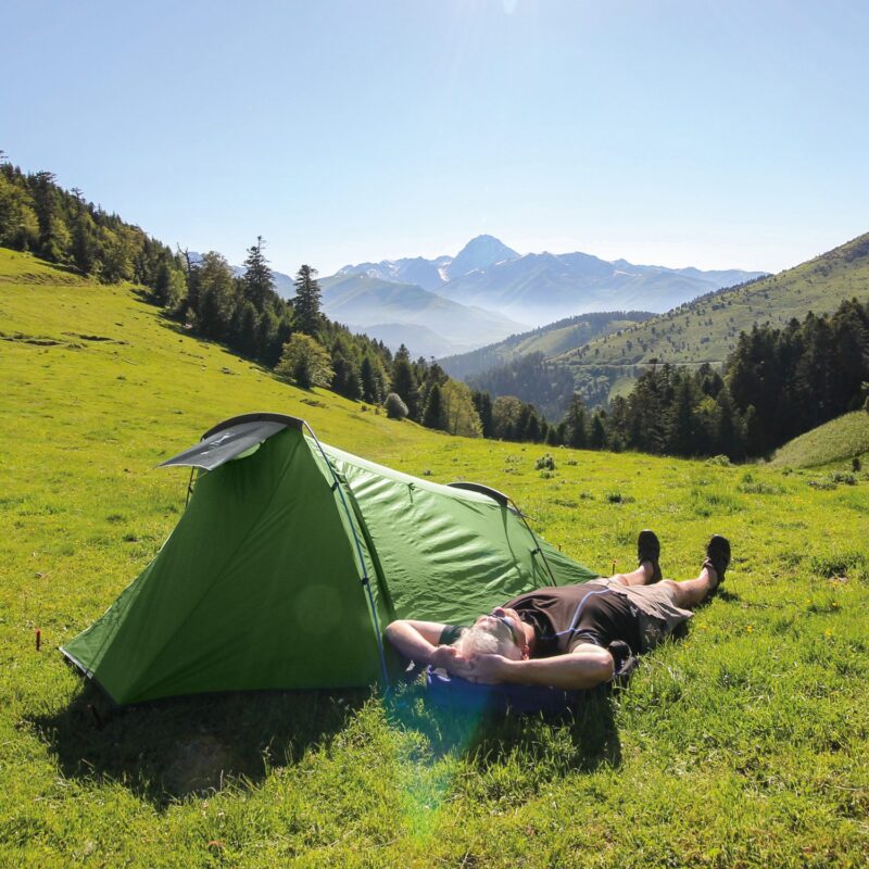 Person lying on their back with hands behind their head beside a bright green tent on a sunlit grassy slope, with tree-lined hills and distant snow-capped mountains under a clear blue sky