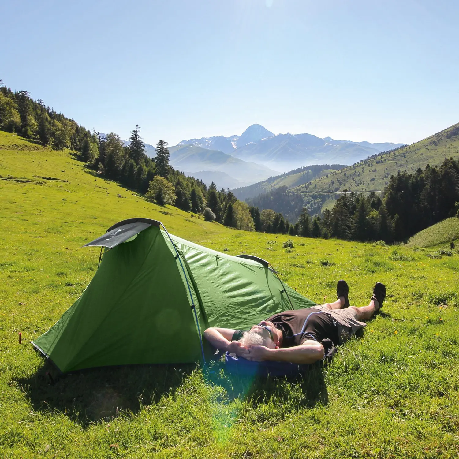 Person lying on their back with hands behind their head beside a bright green tent on a sunlit grassy slope, with tree-lined hills and distant snow-capped mountains under a clear blue sky