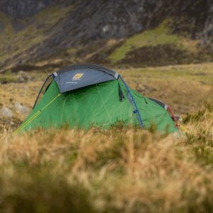 Green dome tent with a grey rainfly, blue and red poles and yellow guylines pitched among windswept grasses in a remote rocky valley with steep grassy hills in the background.
