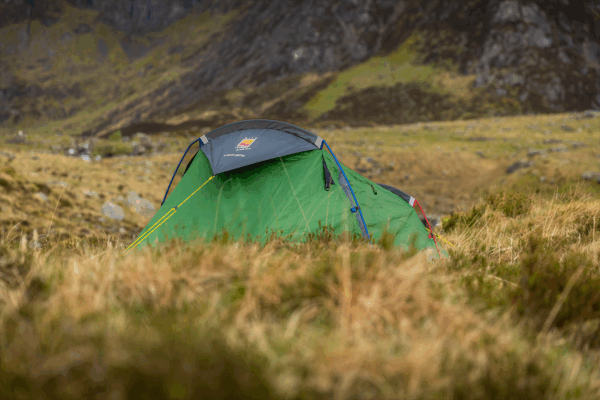 Green dome tent with a grey rainfly, blue and red poles and yellow guylines pitched among windswept grasses in a remote rocky valley with steep grassy hills in the background.