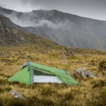 A small green backpacking tent staked on grassy, rocky moorland with yellow guy lines, set against steep grey cliffs and low clouds drifting across the mountains.
