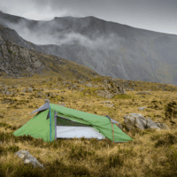 A small green backpacking tent staked on grassy, rocky moorland with yellow guy lines, set against steep grey cliffs and low clouds drifting across the mountains.
