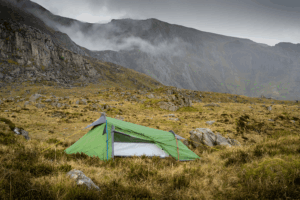 A small green backpacking tent staked on grassy, rocky moorland with yellow guy lines, set against steep grey cliffs and low clouds drifting across the mountains.