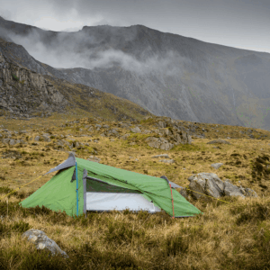 A small green backpacking tent staked on grassy, rocky moorland with yellow guy lines, set against steep grey cliffs and low clouds drifting across the mountains.