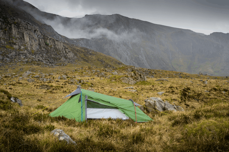 A small green backpacking tent staked on grassy, rocky moorland with yellow guy lines, set against steep grey cliffs and low clouds drifting across the mountains.