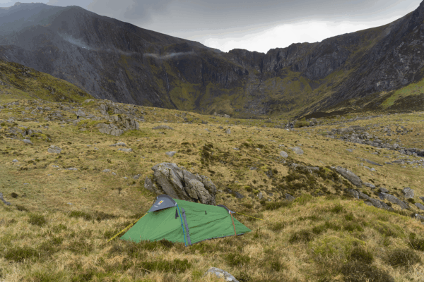 Green tent pitched on grassy, rocky moorland in a wide mountain valley surrounded by steep, dark cliffs under an overcast sky.