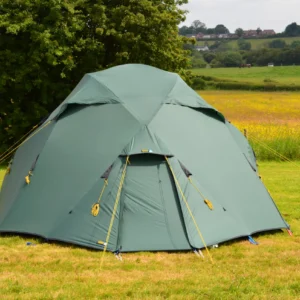 Dome-shaped green camping tent with yellow guy lines staked on a grassy field, backed by a large tree and a distant rolling meadow with houses on the horizon.
