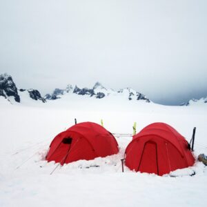 Two bright red dome tents pitched in deep snow on a windswept polar plain, with skis propped nearby and jagged, snow‑covered mountains under a grey overcast sky in the background.