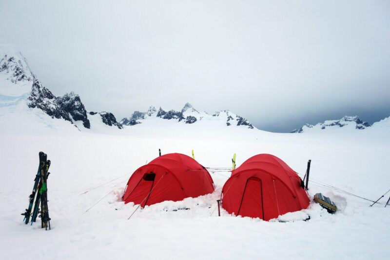 DSC03896enhanced-scaled-4.jpg Two bright red dome tents pitched in deep snow on a windswept polar plain, with skis propped nearby and jagged, snow‑covered mountains under a grey overcast sky in the background.