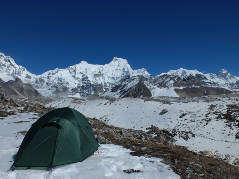 DSCF3897-scaled-4.jpg Green dome tent pitched on a snowy rocky high-altitude plain with expansive snow-covered jagged mountain peaks and glaciers beneath a clear deep-blue sky.