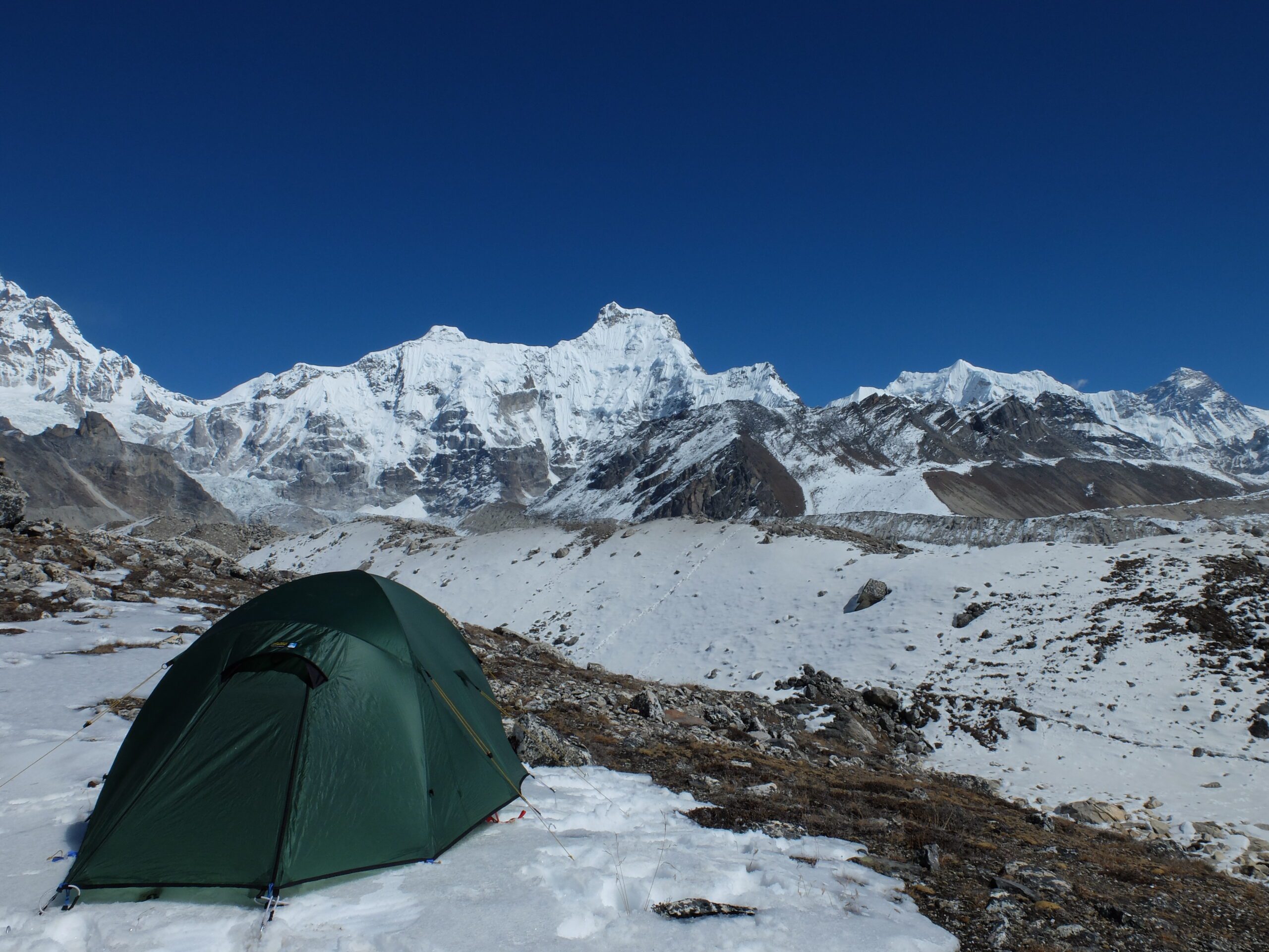 Green dome tent pitched on a snowy rocky high-altitude plain with expansive snow-covered jagged mountain peaks and glaciers beneath a clear deep-blue sky.