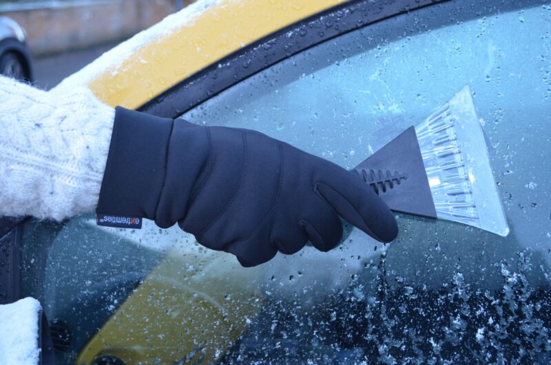 DSC_0645-scaled-2.jpg Black-gloved hand in a cream knit sleeve using a triangular plastic ice scraper to remove frost and ice from a car side window with the yellow car body visible.