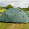 Large green dome tent with yellow guy ropes and triangular vents pitched on a grassy campsite, surrounded by smaller green tents and rolling fields under a cloudy sky.