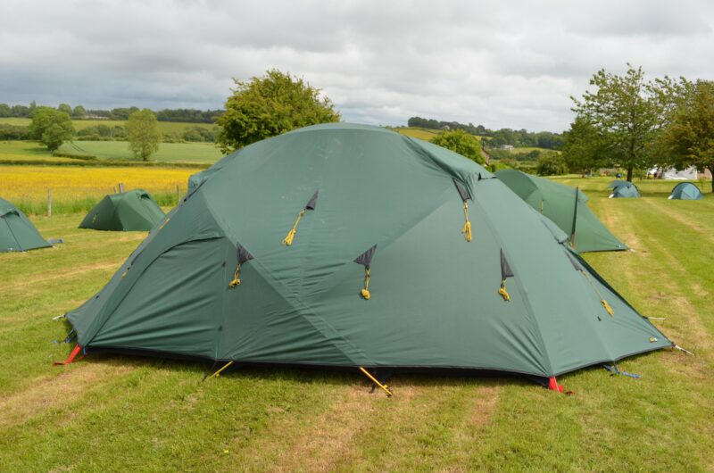 Large green dome tent with yellow guy ropes and triangular vents pitched on a grassy campsite, surrounded by smaller green tents and rolling fields under a cloudy sky.