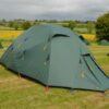 Large green multi‑person tent pitched on a grassy campsite with yellow guy lines and coloured pegs, smaller tents nearby and rolling fields under a cloudy sky in the background.