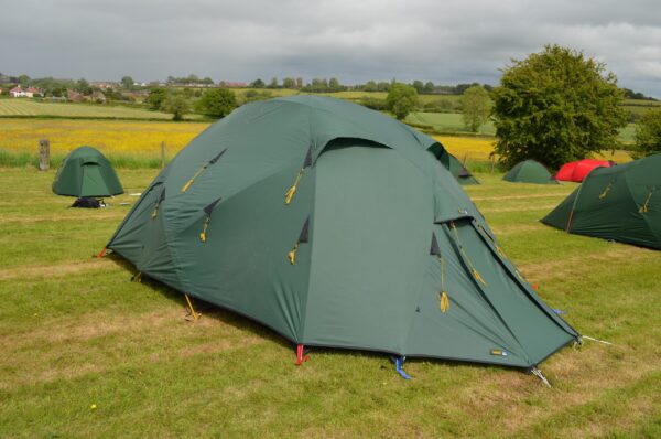 Large green multi‑person tent pitched on a grassy campsite with yellow guy lines and coloured pegs, smaller tents nearby and rolling fields under a cloudy sky in the background.