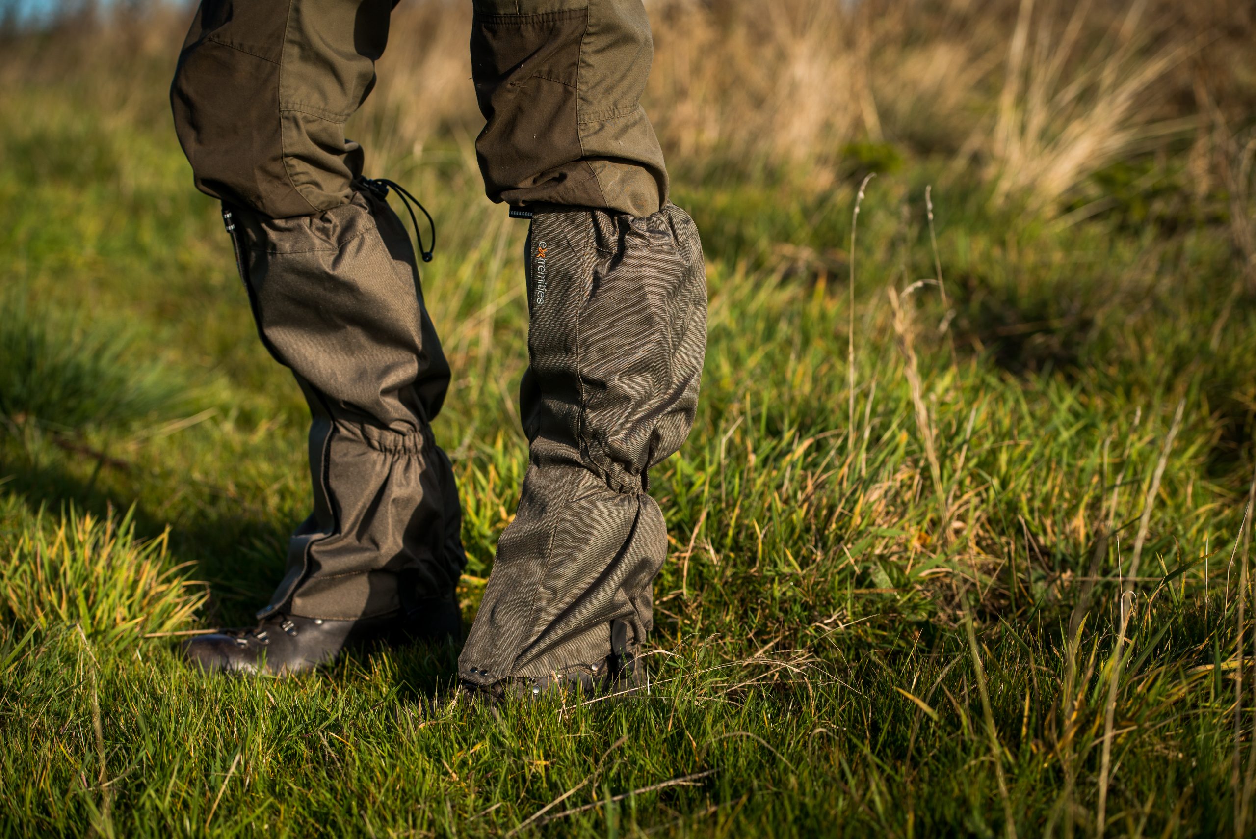 Lower legs and leather hiking boots covered by olive-green gaiters and outdoor trousers standing on a sunlit grassy meadow.