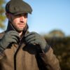 DSC_1919-scaled-4.jpg Bearded man in a tweed coat and flat cap adjusting his collar while wearing olive-green gloves against a sunlit countryside backdrop and clear blue sky