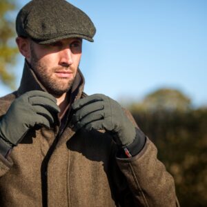 Bearded man in a tweed coat and flat cap adjusting his collar while wearing olive-green gloves against a sunlit countryside backdrop and clear blue sky