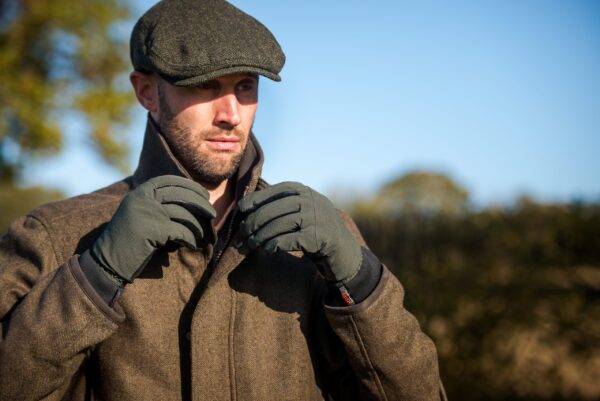 DSC_1919-scaled-4.jpg Bearded man in a tweed coat and flat cap adjusting his collar while wearing olive-green gloves against a sunlit countryside backdrop and clear blue sky