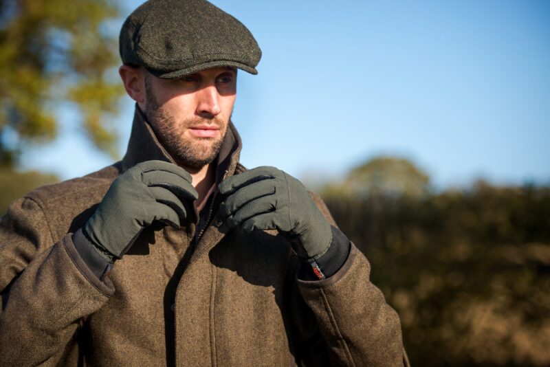 Bearded man in a tweed coat and flat cap adjusting his collar while wearing olive-green gloves against a sunlit countryside backdrop and clear blue sky