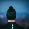 Rear view of a person wearing a black knitted beanie with a pom-pom and white dot pattern and a black jacket with a vertical reflective strip, set against a blurred blue dusk background with soft bokeh lights.