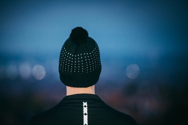 Rear view of a person wearing a black knitted beanie with a pom-pom and white dot pattern and a black jacket with a vertical reflective strip, set against a blurred blue dusk background with soft bokeh lights.