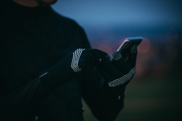 DSC_3871-scaled-2.jpg Close-up of hands wearing black knit touchscreen gloves with white stripe details tapping a smartphone, with a blurred dusk sky and bokeh city lights in the background.