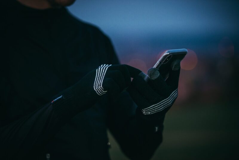 DSC_3871-scaled-2.jpg Close-up of hands wearing black knit touchscreen gloves with white stripe details tapping a smartphone, with a blurred dusk sky and bokeh city lights in the background.
