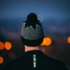 DSC_3914-Low-Res-Copy-Square-1-1.jpg Rear view of a person in a black jacket with a vertical reflective strip, wearing a black-and-white patterned knit beanie with a pompom against a dusk backdrop of blurred orange city lights.