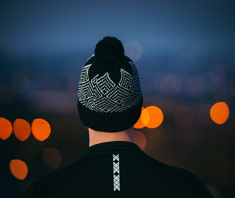 DSC_3914-Low-Res-Copy-Square-1-1.jpg Rear view of a person in a black jacket with a vertical reflective strip, wearing a black-and-white patterned knit beanie with a pompom against a dusk backdrop of blurred orange city lights.