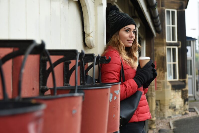DSC_4079-scaled-2.jpg Woman in a red puffer jacket and black beanie leaning against a station-style building, holding a takeaway coffee cup and looking to the right with a row of red metal bins in the foreground.