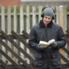 DSC_4239-scaled-2.jpg A man in a grey beanie, over-ear headphones and a black quilted jacket reads a paperback book with gloved hands while standing in front of a wooden fence with diagonal slats.