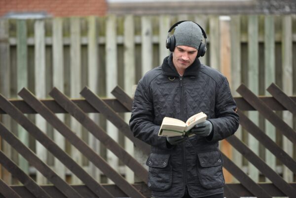 DSC_4239-scaled-2.jpg A man in a grey beanie, over-ear headphones and a black quilted jacket reads a paperback book with gloved hands while standing in front of a wooden fence with diagonal slats.