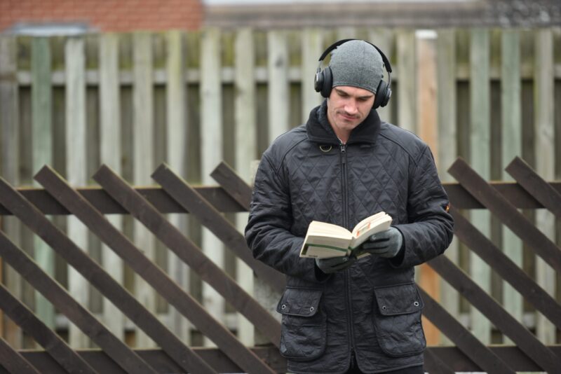 A man in a grey beanie, over-ear headphones and a black quilted jacket reads a paperback book with gloved hands while standing in front of a wooden fence with diagonal slats.