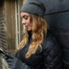 DSC_4340-scaled-4.jpg Woman in a grey beanie and black quilted jacket leans against a dark wooden wall, wearing grey gloves and looking down at her smartphone, her wavy blonde hair falling over her shoulder with a brick building and window behind her.