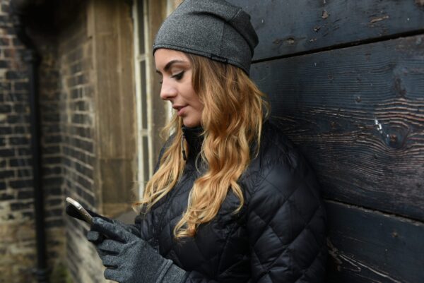 DSC_4340-scaled-4.jpg Woman in a grey beanie and black quilted jacket leans against a dark wooden wall, wearing grey gloves and looking down at her smartphone, her wavy blonde hair falling over her shoulder with a brick building and window behind her.