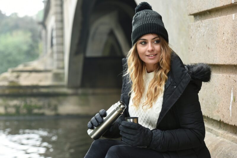 Young woman in a black knit hat, quilted gloves and fur-trimmed coat sits by a stone riverside under a bridge, pouring a hot drink from a stainless-steel thermos into a cup.