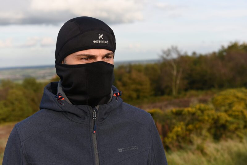 DSC_5133-scaled-2.jpg Person outdoors wearing a black thermal cap with an 'extremities' logo and a matching face mask covering the lower face, dressed in a zipped dark grey jacket against a blurred grassy and wooded landscape.