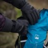 Close-up of a person wearing dark grey insulated gloves and a purple jacket sleeve as they open a bright blue hiking backpack with visible straps and logo against a blurred outdoor background.
