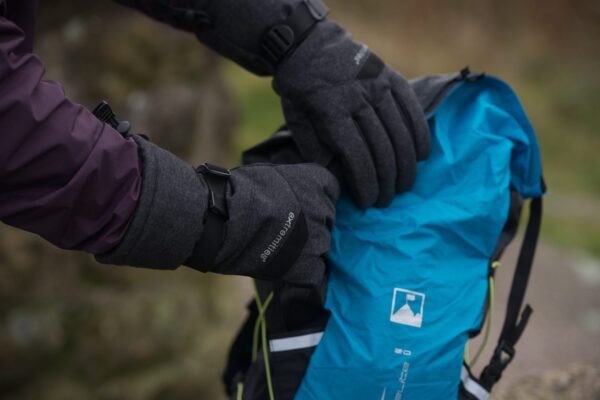Close-up of a person wearing dark grey insulated gloves and a purple jacket sleeve as they open a bright blue hiking backpack with visible straps and logo against a blurred outdoor background.
