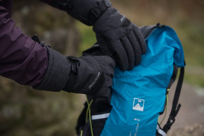 DSC_5313-1.jpg Close-up of a person wearing dark grey insulated gloves and a purple jacket sleeve as they open a bright blue hiking backpack with visible straps and logo against a blurred outdoor background.