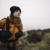 Defence-gloves-1-3.jpg A hiker in a mustard-yellow jacket, black beanie and gloves adjusts his backpack while looking ahead on a foggy moorland trail strewn with rocks.