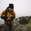 Defence-gloves-2-3.jpg A hiker in a mustard-yellow jacket, black beanie and gloves adjusts his backpack while climbing a narrow rocky path on a foggy grassy hillside.