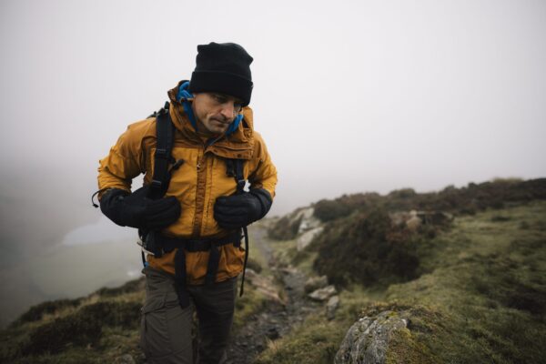 Defence-gloves-2-3.jpg A hiker in a mustard-yellow jacket, black beanie and gloves adjusts his backpack while climbing a narrow rocky path on a foggy grassy hillside.