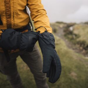 Person in a mustard-yellow jacket and brown trousers fastening black insulated gloves while standing on a misty grassy hillside.